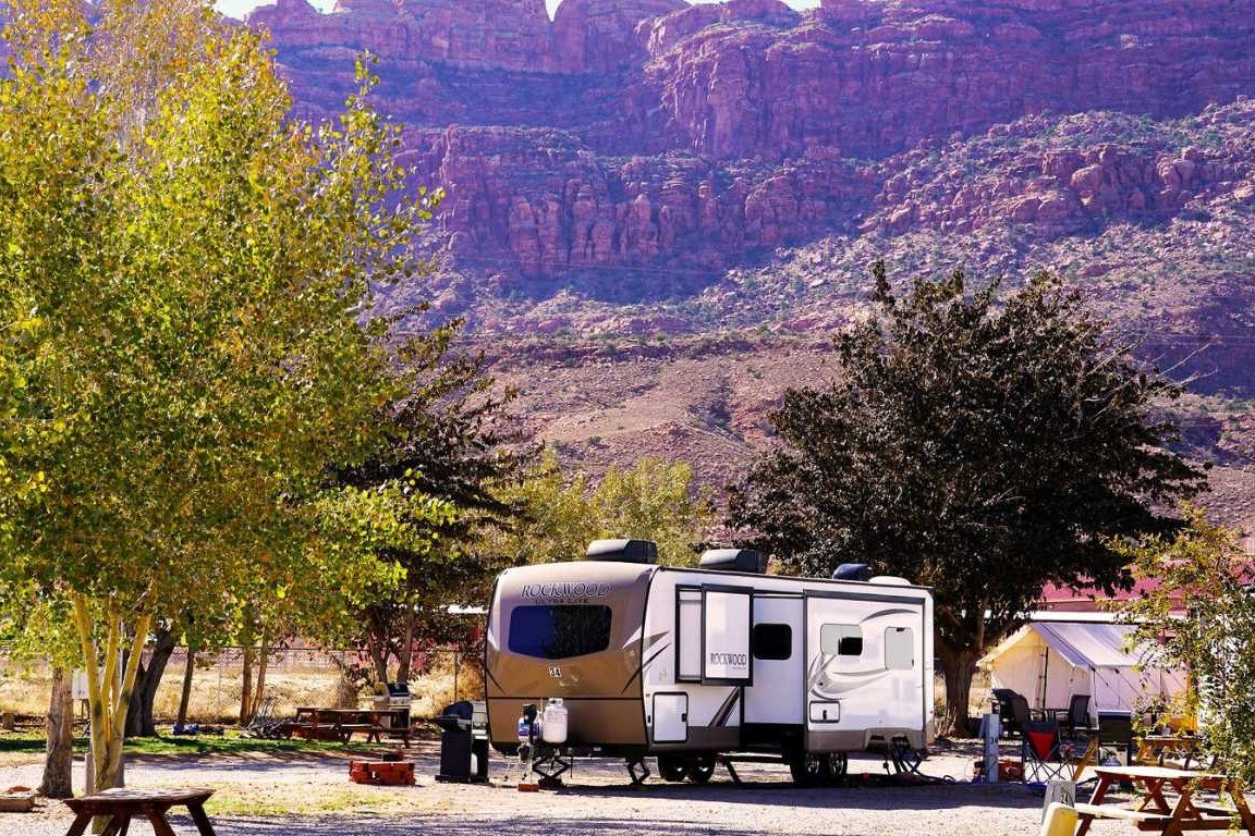 A rental RV under gold cottonwoods with red rock cliffs behind at Moab RV Park